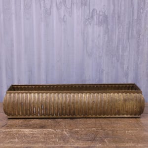 May include: A rectangular brass planter featuring a continuous vertical fluted pattern along its exterior sides. The metal container has a weathered, aged finish with a warm golden-brown tone and a flat, open top design for holding various items.