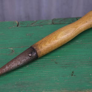 May include: A hand tool featuring a smooth, light-coloured wooden handle attached to a tapered, pointed metal tip. The metal component shows signs of oxidation and wear, while the wooden handle has a natural grain and a rounded, ergonomic end.