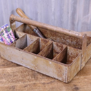 May include: A rectangular metal tool caddy with a central handle and multiple interior compartments, featuring a weathered, rusted finish. A wooden-handled tool rests inside, alongside two small booklets with visible text.