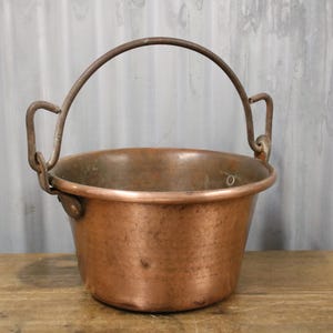 May include: A copper bucket with a curved handle. The bucket has a rounded body and a wide rim. The handle is made of a darker metal and is attached to the bucket with small loops. The bucket is sitting on a wooden surface.