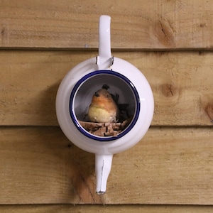 May include: A white teapot-shaped birdhouse with a blue rim and handle, fixed to a wooden wall. The birdhouse contains a small bird and nesting material. This decorative item is designed for outdoor use and adds a touch of whimsy.