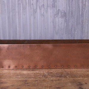 May include: A rectangular copper planter box featuring a smooth, metallic finish with visible vertical and horizontal seams reinforced by small, evenly spaced metal rivets along the edges and the base, creating a structured, industrial appearance.