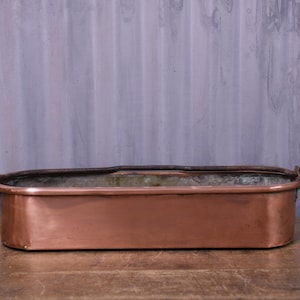 May include: An elongated, rectangular copper planter with rounded corners and a metallic finish. The vessel features a dark, weathered metal rim and two small, integrated handles on the short sides. The interior shows a dull, aged patina.