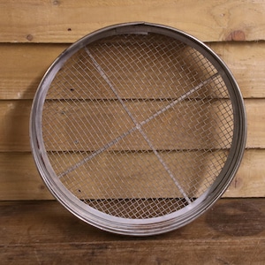 May include: A circular metal sieve featuring a fine wire mesh screen held within a sturdy rim. The frame includes two intersecting metal support bars that cross in the centre to reinforce the structure of the mesh, creating a functional tool.
