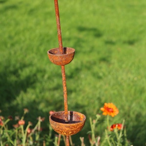 May include: A rusty metal garden sculpture with three bowls stacked on a tall stem. The sculpture is in a garden setting with green grass and orange flowers.