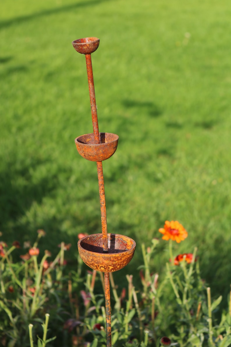 May include: A rusty metal garden sculpture with three bowls stacked on a tall stem. The sculpture is in a garden setting with green grass and orange flowers.