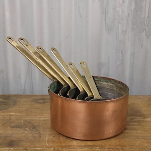 May include: A copper cooking pot with a set of brass measuring ladles. The pot has a rounded shape and a tarnished patina. The ladles are arranged inside the pot, with their handles extending upwards. The background is a weathered, light grey wall.