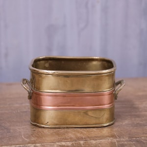May include: A rectangular brass container with rounded corners and a horizontal copper band running across the centre. The vessel features two small metal handles on opposite sides and has a polished metallic finish with visible surface reflections.