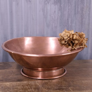 May include: A polished copper bowl with a wide, shallow basin and a circular pedestal base. The bowl features a smooth, reflective metallic surface and contains a cluster of dried, light brown hydrangea flowers resting inside the rim.