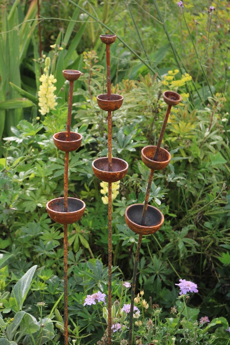 May include: A rusty metal garden sculpture with multiple tiers of bowls. The sculpture is surrounded by green plants and purple flowers.