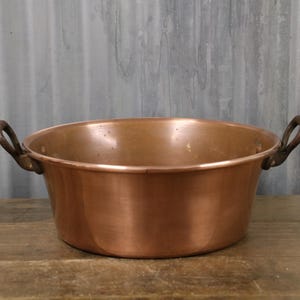 May include: A copper bowl with two curved handles. The bowl has a wide, shallow design and a polished finish. The handles are a darker colour, possibly bronze or iron. The bowl sits on a wooden surface.