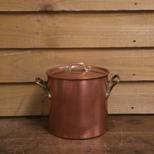 May include: A cylindrical copper stockpot with a matching lid and two brass side handles. The pot features a polished metallic finish with a warm reddish-brown hue. The lid includes a small brass handle centred on top for easy removal.