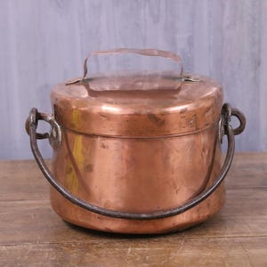 May include: A round copper container with a flat lid and a small metal handle on top. The container features a larger, curved iron swing handle attached to the sides with metal loops. The copper surface shows a warm, metallic finish with signs of wear.