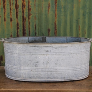 May include: An oval galvanised metal wash tub with a weathered silver-grey finish. The basin features horizontal ribbed detailing along the exterior walls, two side handles, and a faint circular stamped emblem centred on the front surface.