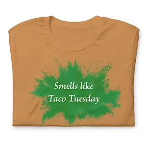 May include: Mustard-colored t-shirt with the text "Smells like Taco Tuesday" in white letters over a green splash design. The shirt is folded, showing the neckline and the front of the garment.
