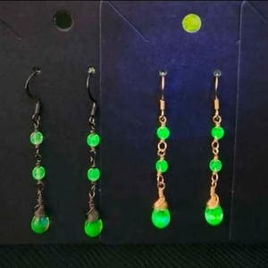 May include: A collection of handmade earrings with a glowing green bead design. The earrings feature various metal finishes, including copper, black, and gold, with a dangling teardrop-shaped bead at the bottom. The earrings are displayed on a black background.