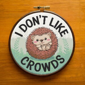 Peut inclure: Art de broderie sur un cercle avec un fond vert et un mignon hérisson de dessin animé. Le texte "Je n'aime pas les foules" est écrit en noir autour du hérisson.