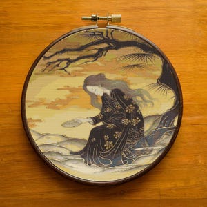 Op de afbeelding: Een ronde borduurring met een kruissteekontwerp van een vrouw in een zwarte kimono met gouden bloemaccenten, zittend onder een boom. De achtergrond heeft een gele en oranje gradiënt, met een donkerbruin houten frame.