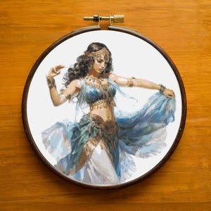Puede incluir: Bordado de punto de cruz de una mujer con un traje azul y blanco fluido, bailando. El bordado está colocado en un aro de madera redondo. La mujer tiene el pelo oscuro y está adornada con joyas. El fondo es blanco.