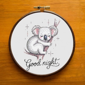 May include: A cross-stitch embroidery in a wooden hoop features a sleeping koala bear on a branch. The koala is white and gray, with closed eyes. The words "Good night" are written in cursive below the koala. The background is white.
