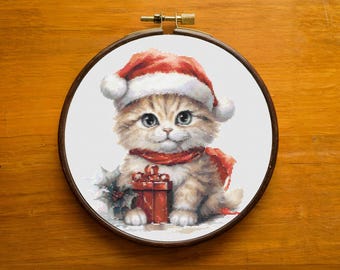 May include: Cross-stitch embroidery of a kitten wearing a Santa hat and scarf, holding a red gift box. The kitten has light brown fur and blue eyes. The embroidery is set in a wooden hoop.