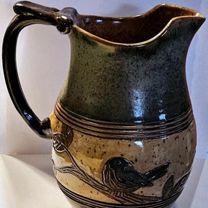 May include: A brown and green ceramic pitcher with a bird design. The pitcher has a wide mouth and a handle.