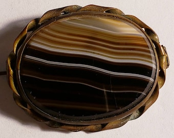 May include: Oval-shaped vintage brooch with a polished agate stone. The stone features bands of brown, white, and black. The brooch has a decorative, twisted metal frame in a gold tone, and a pin clasp on the back.