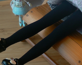 May include: A pair of black tights with a decorative button on the skirt. The tights are worn by a doll sitting on a wooden chair.