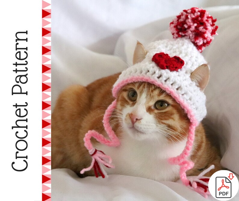 May include: A white crocheted cat hat with a pink brim and a red heart appliqu&eacute;. The hat has a pink pom-pom on top and a long pink tassel hanging from the side. The hat is worn by a cat with orange and white fur.