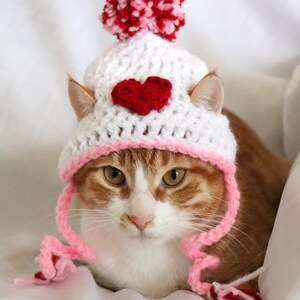May include: A white and pink crocheted cat hat with a red heart on the front and a pink and red pom-pom on top. The hat is worn by a cat with orange and white fur.
