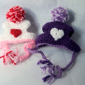 May include: Two crocheted hats with heart-shaped embellishments and pom-poms. One hat is white with a pink trim and a red heart, the other is purple with a white heart.