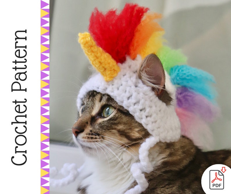May include: A cat wearing a white crocheted unicorn hat with a rainbow mane. The hat has a yellow horn and a white crocheted chin strap.