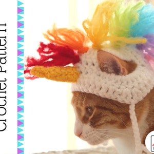 May include: A white crocheted cat hat with a rainbow mane and a yellow horn. The hat is on a cat with orange fur.