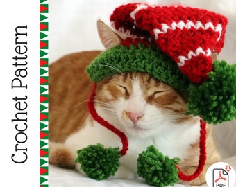 Può includere: Un cappello da elfo all'uncinetto con filato verde e rosso e pon pon bianchi. Il cappello è sulla testa di un gatto.