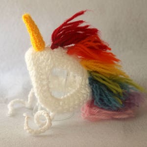 May include: White crocheted unicorn head with a yellow horn and a rainbow mane. The mane is made of different colored yarn, including red, orange, yellow, green, blue, and pink.
