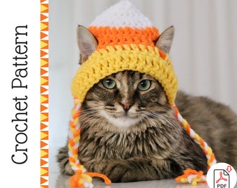 May include: A brown tabby cat wearing a crocheted candy corn hat. The hat is white, orange, and yellow. The cat is looking at the camera.