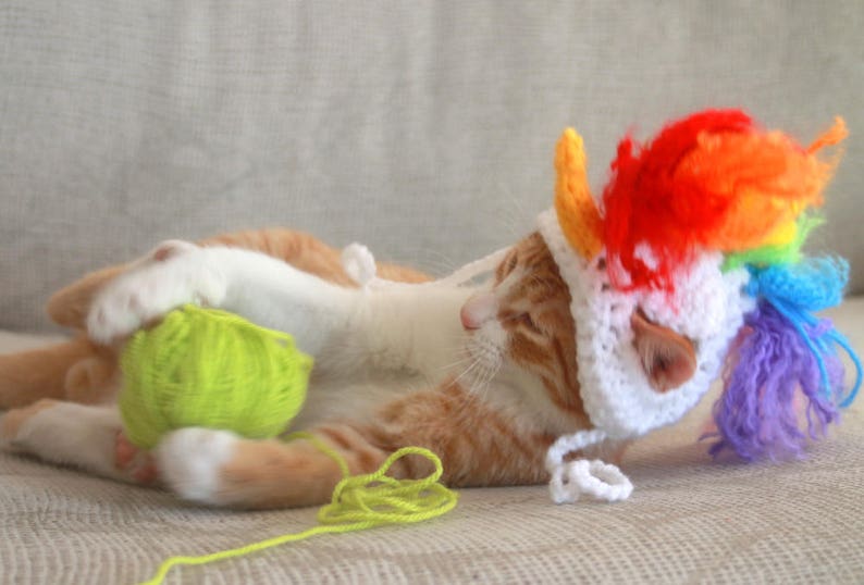 May include: A ginger cat wearing a white crocheted unicorn hat with a rainbow mane and tail. The cat is playing with a green yarn ball.