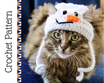May include: A brown tabby cat wearing a white crocheted snowman hat with an orange carrot nose. The cat is looking directly at the camera.