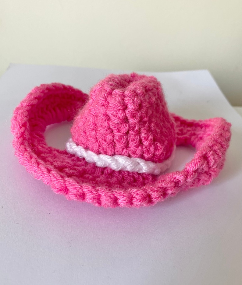 K&ouml;nnte beinhalten: Ein rosa geh&auml;kelter Cowboyhut mit einem wei&szlig;en geh&auml;kelten Band um die Krone.