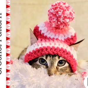 May include: A tabby cat wearing a handmade crochet hat with a pink and white striped pattern and a matching pom-pom. The hat is labeled "Crochet Pattern" in white text on the left side of the image. A PDF download icon is in the bottom right corner.