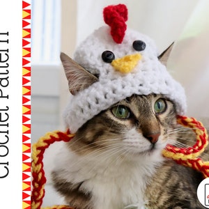 May include: A white crocheted chicken hat with red and yellow accents, worn by a tabby cat. The hat has black button eyes and a yellow beak. The cat is looking to the right of the image.