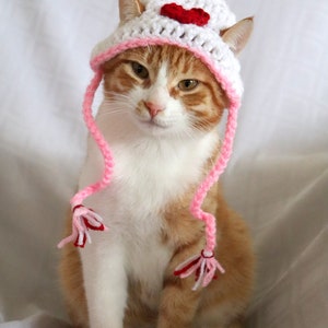 May include: A white and pink crocheted cat hat with a red heart and a pink pom-pom on top. The hat has long pink ties with white tassels.