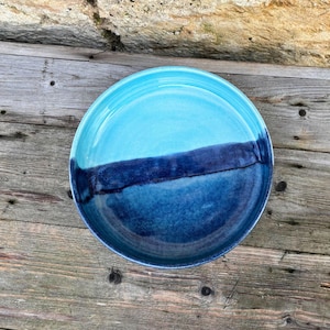 Pode incluir: Uma tigela redonda de cerâmica com um gradiente de tons de azul. A metade superior é turquesa claro, passando para uma faixa azul mais escura e, em seguida, um azul marinho profundo na parte inferior. A tigela está sobre uma superfície de madeira.