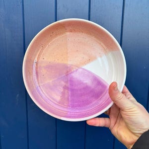 Pode incluir: Um prato de cerâmica feito à mão com um esquema de cores único. O prato é dividido em quadrantes de pêssego, branco e tons de rosa e roxo. O prato tem uma borda ligeiramente elevada e uma textura salpicada.