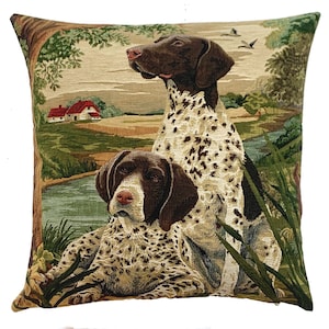 Könnte beinhalten: Quadratisches Zierkissen mit einem Wandteppichmuster, das zwei Deutsch Kurzhaar Hunde in einer Naturlandschaft zeigt. Die Hunde haben braun-weiß geflecktes Fell. Der Hintergrund umfasst einen Fluss, Bäume und ein Haus.