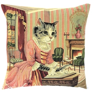 May include: A square throw cushion featuring a woven tapestry design of a tabby cat dressed in a pink gown, sitting at a desk and writing with a quill. The background depicts a room with striped wallpaper, a fireplace, and a window with red curtains.