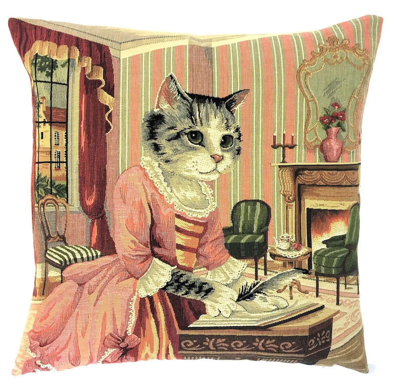 May include: A square throw cushion featuring a woven tapestry design of a tabby cat dressed in a pink gown, sitting at a desk and writing with a quill. The background depicts a room with striped wallpaper, a fireplace, and a window with red curtains.
