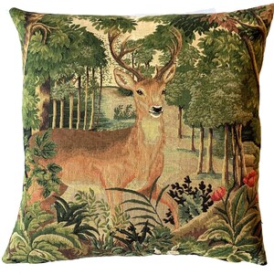 May include: A tapestry pillow featuring a brown deer with antlers standing in a green forest. The deer is surrounded by green trees and bushes. The pillow is made of a soft, woven fabric.
