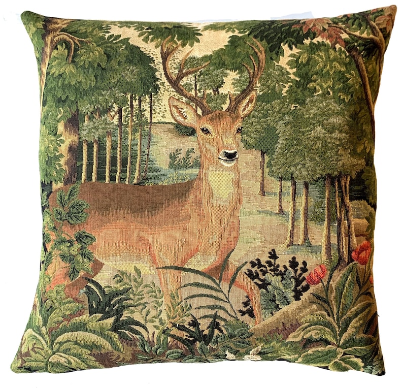 May include: A tapestry pillow featuring a brown deer with antlers standing in a green forest. The deer is surrounded by green trees and bushes. The pillow is made of a soft, woven fabric.