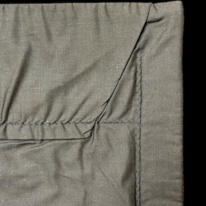 May include: A dark gray fabric textile featuring a smooth, flat border edge joined to a gathered, pleated section. The seam connecting the two parts is reinforced with visible, thick stitching that runs horizontally across the fabric surface.